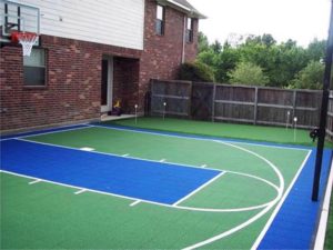 Green and blue, in-home basketball court with a fence and brick wall. 